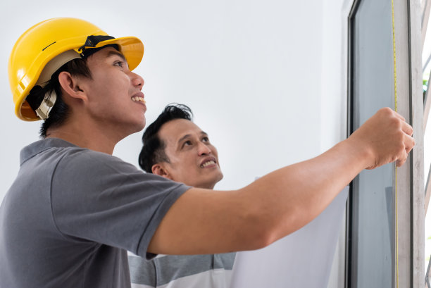 戴着安全帽的工头在进行一项新的家庭改造工程时，用卷尺测量窗框。图片下载