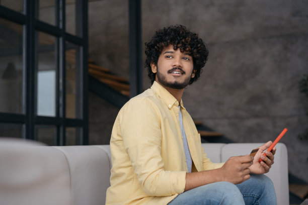 微笑的印度男子拿着信用卡在网上用手机购物。移动银行。英俊的亚洲男人在网上订餐，看向别处，坐在家里，复印空间图片下载