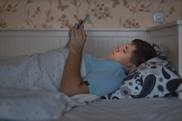 白种男性疲惫地躺在床上上网。手机依赖，网络陷阱，社交媒体成瘾。图片下载