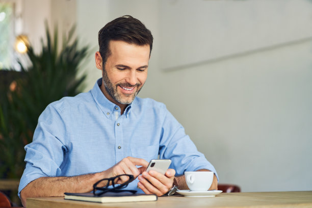 英俊的成年男子坐在桌子旁，在咖啡厅用智能手机图片下载