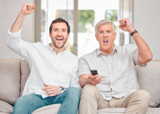 一个英俊的年轻男子和他的老父亲在家里的沙发上为他们的球队加油图片下载