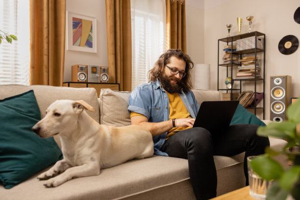一位满脸笑容、戴着眼镜的大胡子男子在他心爱的狗狗陪伴下远程工作。这个人用他的笔记本电脑，浏览他喜欢的网站，给同事发电子邮件图片下载