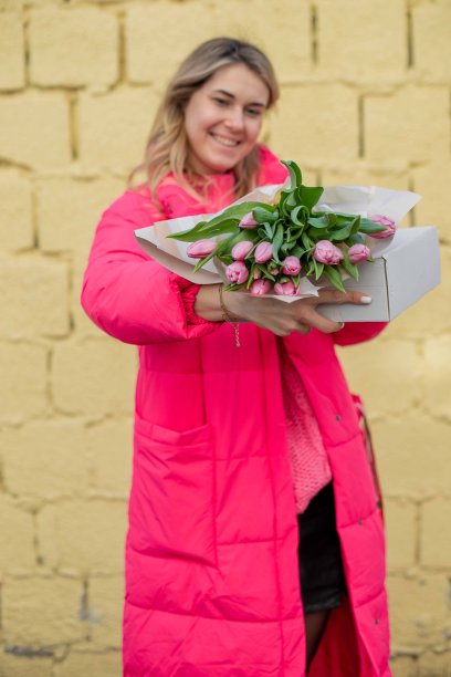 垂直照片平静害羞微笑的金发女人粉红色的夹克，拿着大的鲜花，郁金香。家庭意外图片下载