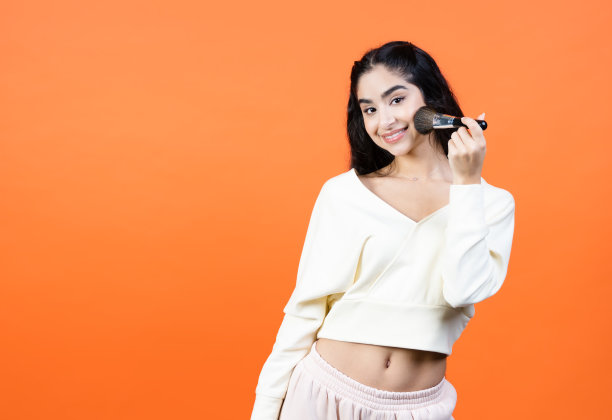 美丽的黑发女人与化妆刷在她的手与复制空间的橙色墙壁上。广告背景与复制空间。图片下载