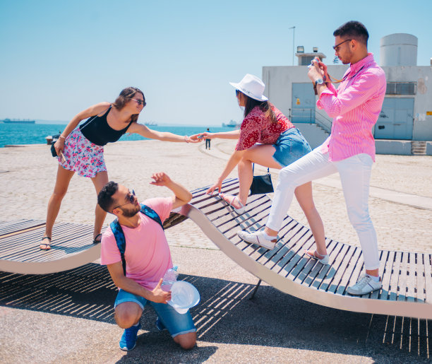 在一个阳光明媚的日子里，四个游客朋友在一条现代抽象的长凳上散步，享受着拍照的乐趣。欣赏湛蓝碧绿的大海。图片下载