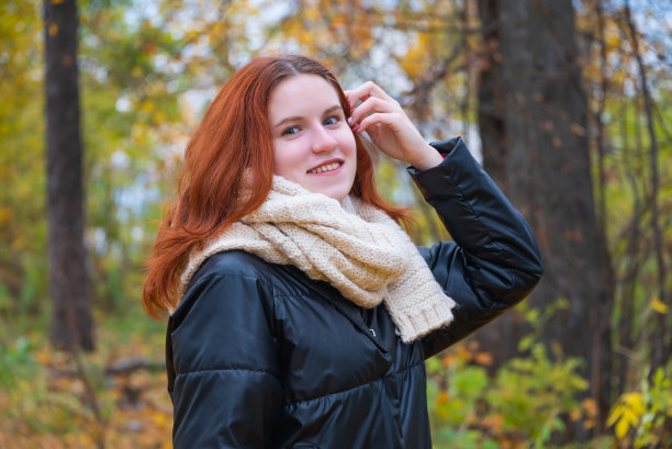一个穿着夹克的红头发微笑女孩的肖像，她用手拉直了头发。以秋的自然为背景，以人的情感为概念图片下载