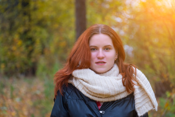 一个红发女孩的肖像，穿着夹克和围巾，背景是秋天的自然和明亮的太阳概念的人类情感图片下载