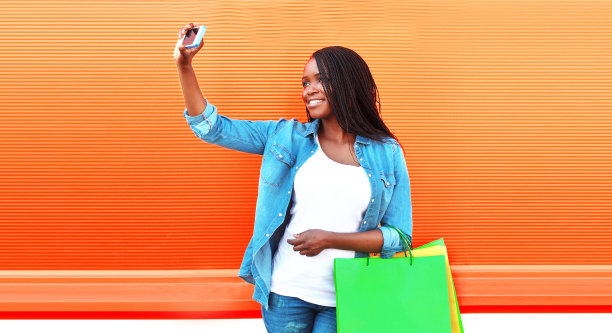 快乐微笑的非洲妇女拿着购物袋在城市街道上用智能手机自拍图片下载