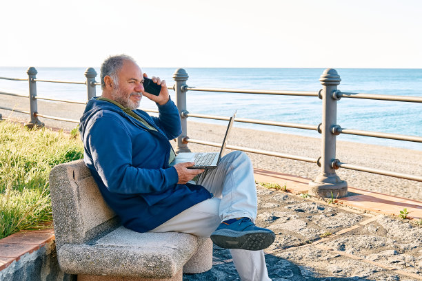 一个大胡子的成熟男人坐在海边的长椅上，带着笔记本电脑在户外工作。现代生活方式，联系，商业，自由职业的概念。一个面带微笑的白发男人在用智能手机聊天图片下载