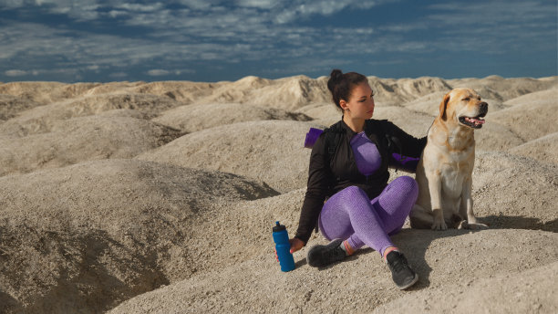 女子在运动紫色的背包坐在黄色的沙丘上与狗拉布拉多寻回犬看向远方在阳光明媚的一天图片下载