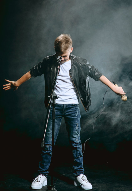 穿着白色t恤、蓝色牛仔裤和皮夹克的白人少年的肖像，麦克风在黑暗的背景下唱歌。爱好与荣耀理念图片下载