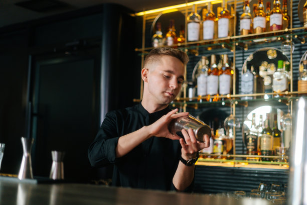 年轻英俊的男调酒师站在吧台后面，用摇酒器调酒。图片下载