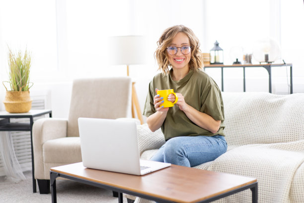 女性眼镜使用笔记本电脑，坐在沙发上的大窗户背景在家里室内图片下载