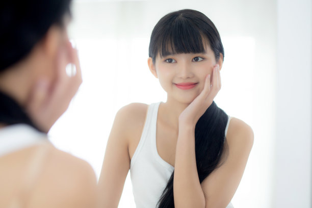 美丽的年轻亚洲女性的脸与快乐的微笑和触摸脸颊与迷人的镜子，化妆美丽的面部女孩与护肤美容治疗，皮肤护理和健康的概念。图片下载