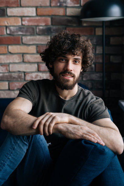 有自信的大胡子年轻人的垂直肖像，微笑着看着镜头，坐在舒适的沙发在客厅的砖墙。图片下载