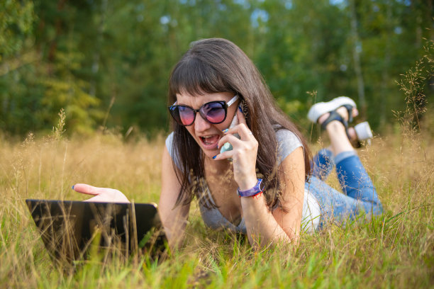 一个年轻女子感情用事地打电话，看着笔记本电脑。女自由职业者在阳光明媚的草地上工作图片下载