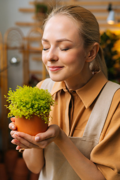 妩媚微笑的女花商的特写脸，围裙上散发着太阳花的香味，手里拿着花盆，在花店里闭着眼睛。图片下载