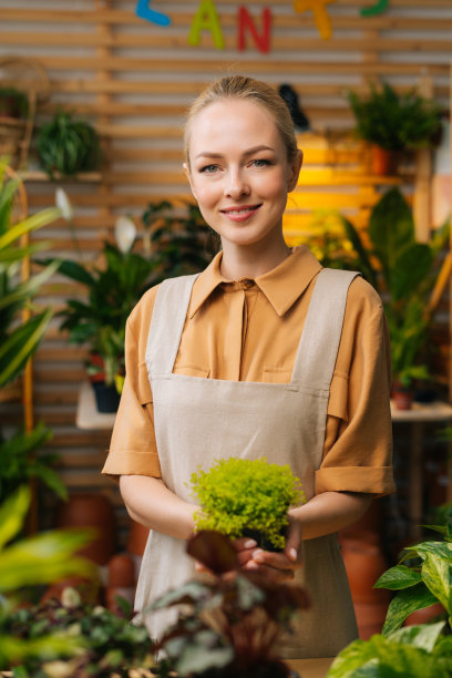 女花商面带微笑，穿着围裙，手里拿着花盆，站在花店里，看着相机。图片下载