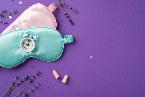 俯视图照片的两个丝绸睡眠面具粉红色和蓝色的一个小白色时钟在它和分散的五彩纸屑形状的星星与薰衣草的树枝在紫色孤立的背景copyspace图片下载