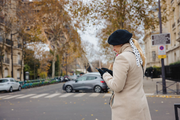 穿着外套的金发女人站在巴黎的人行道上，看着她的手机叫出租车图片下载