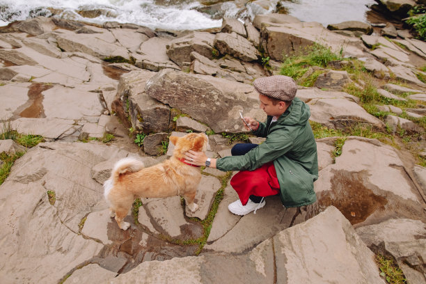 时尚的旅行者喜欢这种景色，他和他的狗坐在山上的一条河边，用手机拍下了照片。旅行和流浪的概念。图片下载