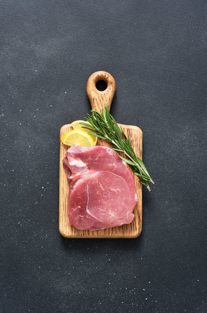 新鲜的生肉放在木板上，迷迭香和柠檬放在黑色的背景上，猪肉图片下载