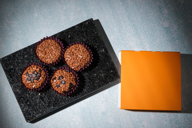 低调照片。Brigadeiros排列在一块闪亮的黑色大理石上，旁边是一个橙色的盒子顶视图。图片下载