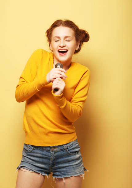 穿着短裤和黄色毛衣的红头发年轻女子兴高采烈地拿着麦克风唱歌图片下载