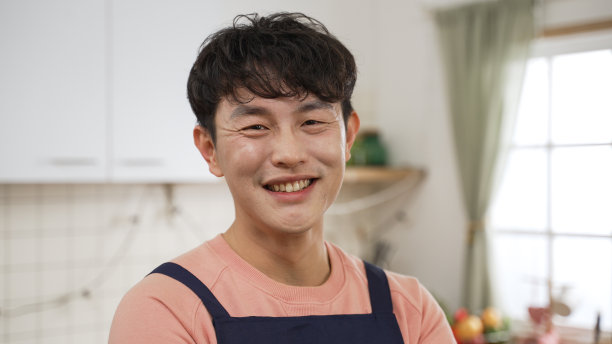 在家里的一间现代明亮的餐厅里，一名穿着围裙的亚裔男子笑容满面地看着镜头，用肩拍的照片图片下载