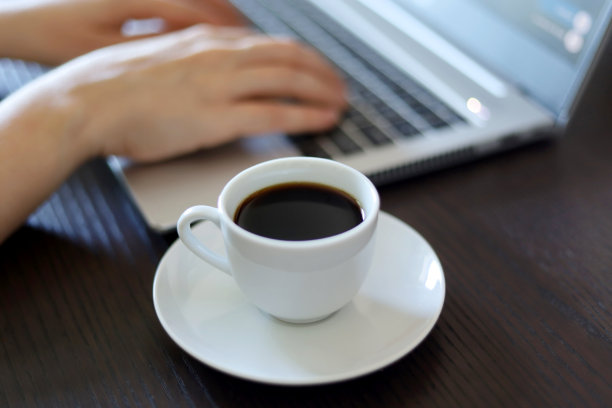 咖啡杯与茶托背景上的女性手在笔记本电脑键盘图片下载