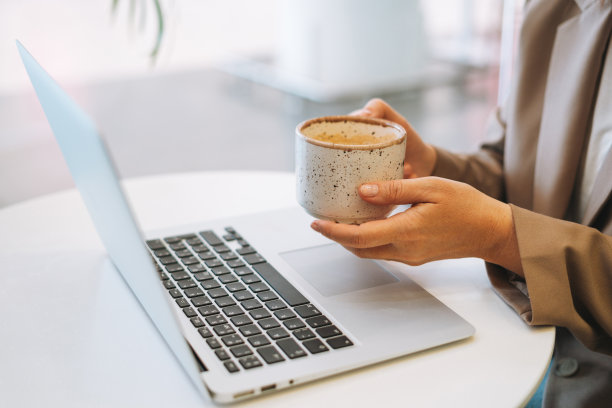 留着长发的商务女性，穿着时尚的米色西装，在桌子上喝咖啡，在笔记本电脑前工作图片下载