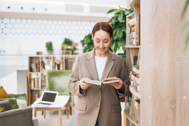 成年人微笑着，棕色头发，四十岁的商业女性，在公共场所，绿色的开放空间办公室，共同工作，穿着时尚的米色西装和牛仔裤。友好的老师或导师在图书馆的书图片下载