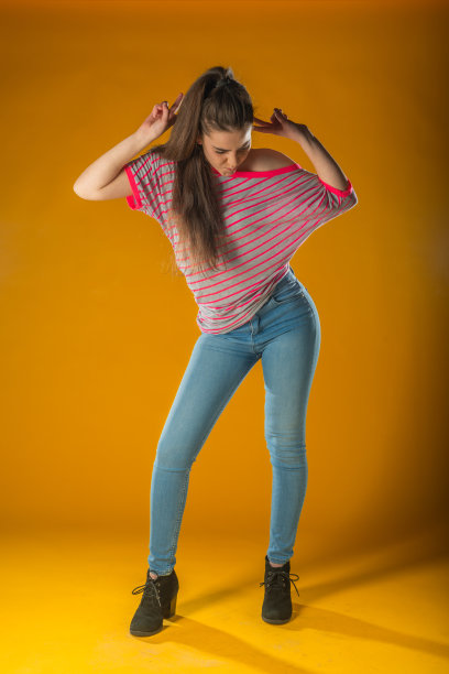 美丽的年轻女子在粉红色的迷你连衣裙摆姿势，手放在臀部，呈现的东西和看向别处。四分之三长度工作室拍摄黄色背景。图片下载