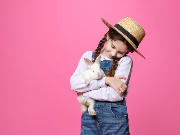 小女孩穿着牛仔裤，戴着草帽，抱着一只小白兔，背景是孤立的粉红色。图片下载