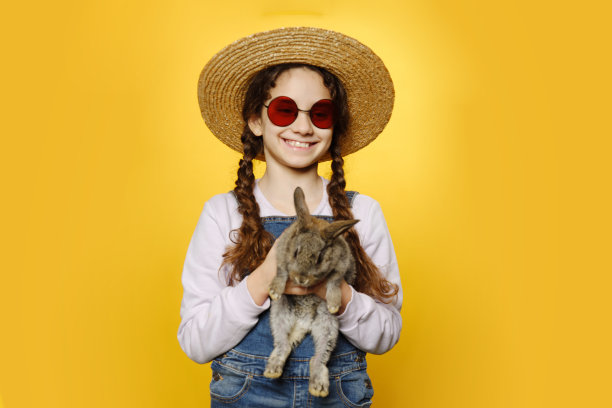 前面是一个穿着牛仔裤的小女孩，戴着草帽和红色太阳镜，手里抱着一只兔子，孤立的黄色背景。图片下载