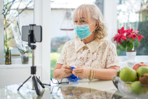 在新冠肺炎疫情期间，一位戴着防护口罩和手套的成熟女性，坐在装有三脚架的智能手机前，与家人视频通话。图片下载