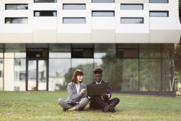 多种族的男女同事坐在办公室外的草地上用笔记本电脑工作图片下载