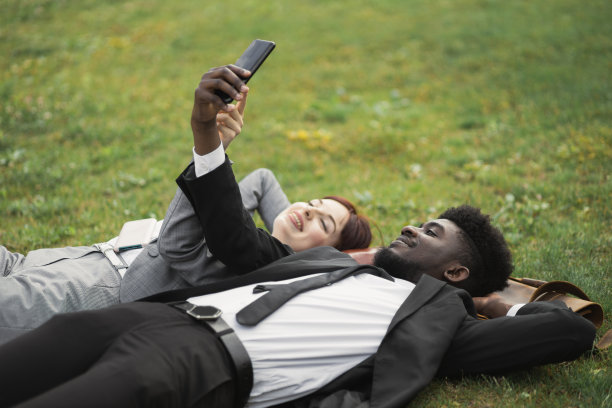 穿着考究的多种族青年商人躺在绿色的草地上，手里拿着智能手机图片下载