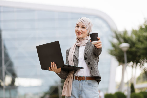 穆斯林妇女穿着时髦的休闲装，站在现代建筑外，用笔记本电脑上网图片下载