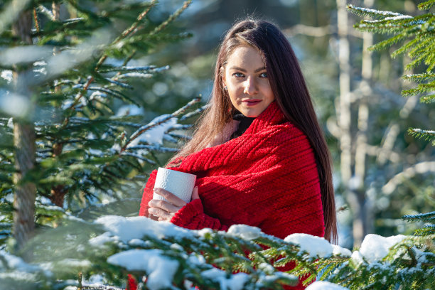 美丽的女人肩上披着红色的格子呢，在冬木里端着一杯茶，画着冬的肖像图片下载