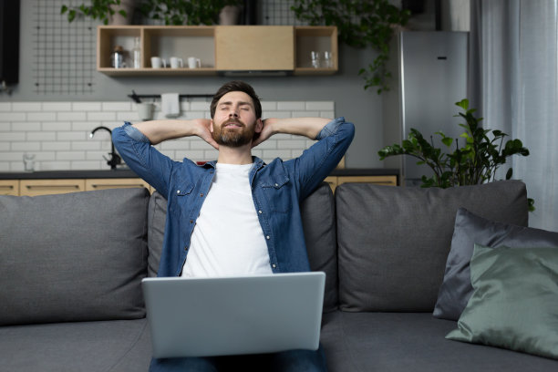 在家的男子坐在沙发上用笔记本电脑工作，双手放在脑后打瞌睡图片下载