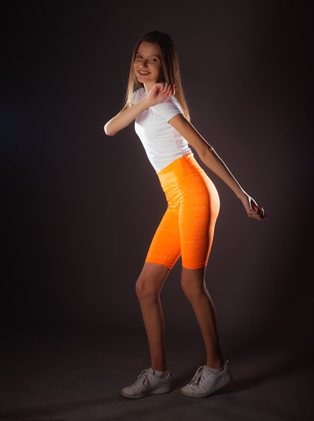 一个微笑的年轻女运动员站在工作室的肖像，穿着白色的上衣和短橙色的打底裤图片下载