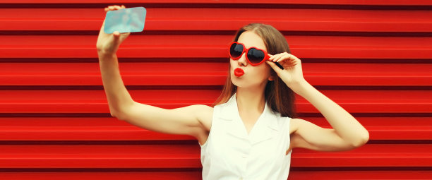 年轻女子的自拍照片，用智能手机吹着嘴唇，涂着红色的口红，在生动的背景上戴着红色的心形太阳镜，送出甜蜜的飞吻图片下载