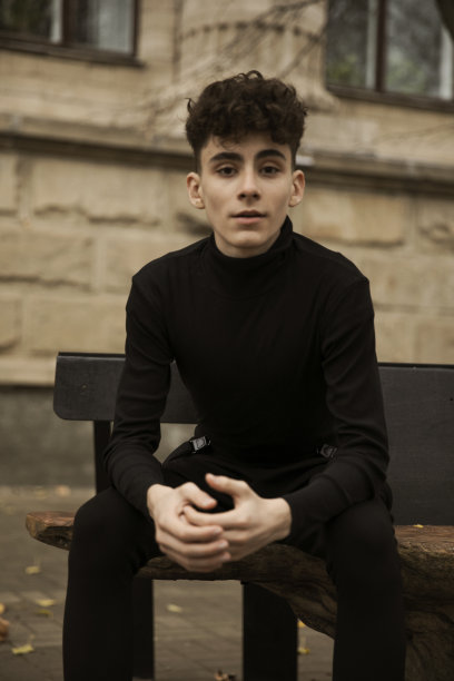 正面照片是一个穿着黑色衣服的年轻男性模特，在一幢建筑后面摆姿势，留着现代发型。图片下载