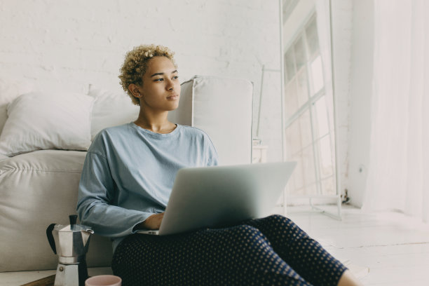 这是一幅现代女性作家坐在地板上沉思的照片，在她舒适的轻简约的公寓里，膝盖上放着笔记本电脑，穿着家装，旁边放着咖啡壶和杯子图片下载