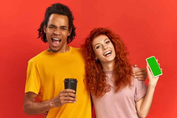 好玩的积极的混合种族夫妇拥抱和快乐地唱歌，提高能量和情绪与智能手机上的酷音乐，喝新鲜的咖啡，看起来乐观和娱乐，在红色的背景。图片下载