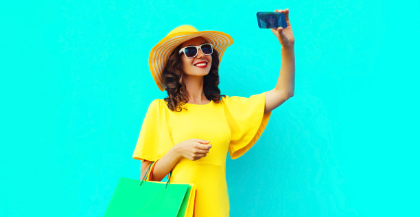 美丽快乐微笑的女人用智能手机与购物袋穿着黄色连衣裙，夏天草帽在彩色蓝色的背景图片下载