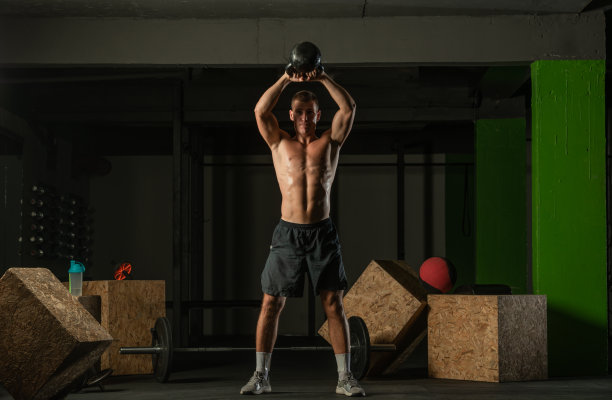 一个英俊的男子赤裸上身，在黑暗的背景下用壶铃运动的全身照片图片下载