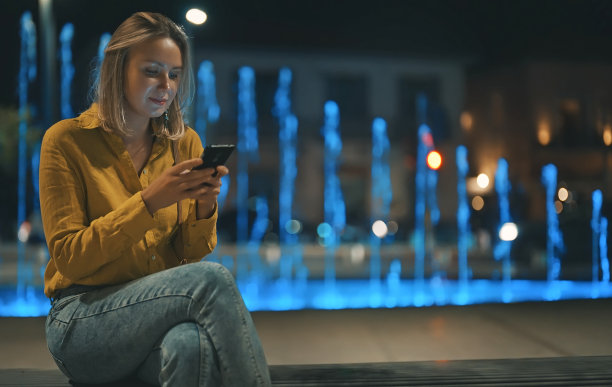 夏日傍晚，手持智能手机的女子坐在喷泉旁。图片下载