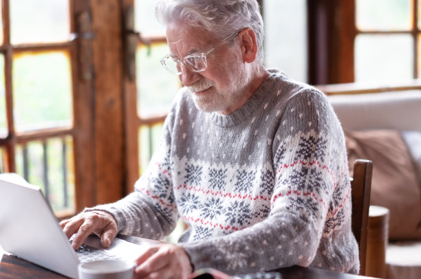满脸胡须的老人穿着毛衣，面带微笑地坐在客厅里，手里拿着咖啡杯，手里拿着笔记本电脑。无忧无虑的老人喜欢科技和社交。林中的乡村小屋图片下载